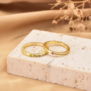 Puede incluir: Dos anillos de oro con pequeñas piedras preciosas. El anillo de la izquierda tiene una piedra preciosa verde y la inscripción "Nicole 10.08". El anillo de la derecha tiene una piedra preciosa rosa y la inscripción "Estoy escuchando".