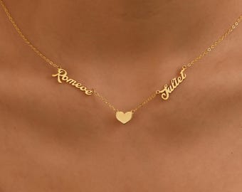 May include: A gold-colored necklace featuring the names "Romeo" and "Juliet" on either side of a small heart pendant. The necklace is worn around the neck and is displayed against a neutral background.