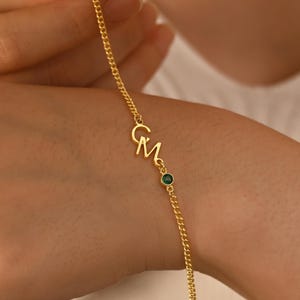 Può includere: Un braccialetto a catena color oro con le iniziali "C M" in corsivo. Una piccola gemma verde è incastonata tra le iniziali e la catena. Il braccialetto è mostrato su un polso.