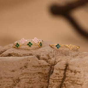 Könnte beinhalten: Zwei goldfarbene Ringe mit kleinen kleeblattförmigen Akzenten. Der linke Ring hat grüne Kleeblattmotive und klare Steine, während der rechte Ring ein blaues Kleeblattmotiv und klare Steine entlang des dünnen Metallbandes aufweist.