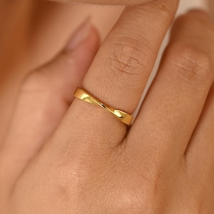 Op de afbeelding: Een goudkleurige ring met een gedraaid ontwerp. De ring wordt aan een vinger gedragen en toont zijn gladde, reflecterende oppervlak. Het sieraad heeft een minimalistische esthetiek, geschikt voor dagelijks gebruik.