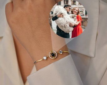 Könnte beinhalten: Ein goldenes Armband mit einem kleinen runden Anhänger, der ein schwarzes Auge in der Mitte hat. Der Anhänger ist von kleinen klaren Steinen umgeben. Das Armband befindet sich am Handgelenk einer Person. Der Anhänger enthält ein verstecktes Bild, das sichtbar wird, wenn man durch das Auge schaut.