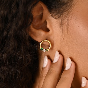 May include: Gold-coloured hoop earrings featuring three small gemstones: green, white, and green. The earrings are circular and worn on an ear. The gemstones are set in a row on the bottom of the hoop.
