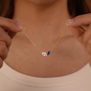 Puede incluir: Un delicado collar de oro con tres piedras preciosas de talla marquesa: una transparente, una azul y una transparente. El collar lo sostiene una persona. La cadena es fina y las piedras están juntas.