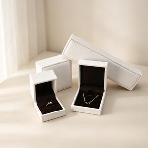 May include: White jewelry boxes with black velvet interiors. One box holds a ring, another a necklace. The boxes are open, displaying the jewelry. A rectangular box and a square box are also present.