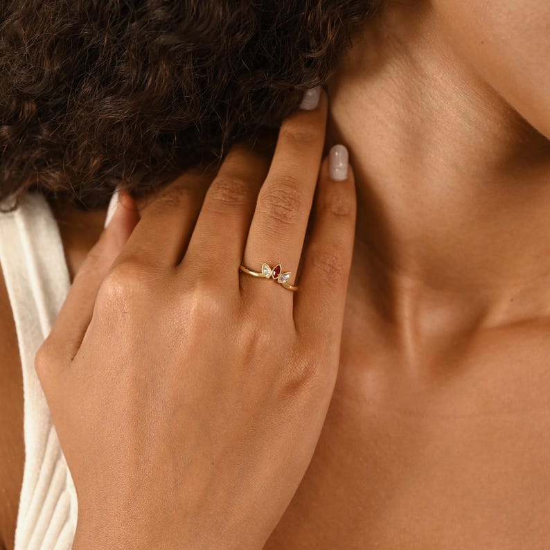 K&ouml;nnte beinhalten: Ein goldener Ring mit einer V-f&ouml;rmigen Ringschiene und drei kleinen Steinen in einer Anordnung. Der mittlere Stein ist ein tiefroter Edelstein im Marquise-Schliff, flankiert von zwei kleineren, klaren Steinen im Marquise-Schliff auf beiden Seiten.