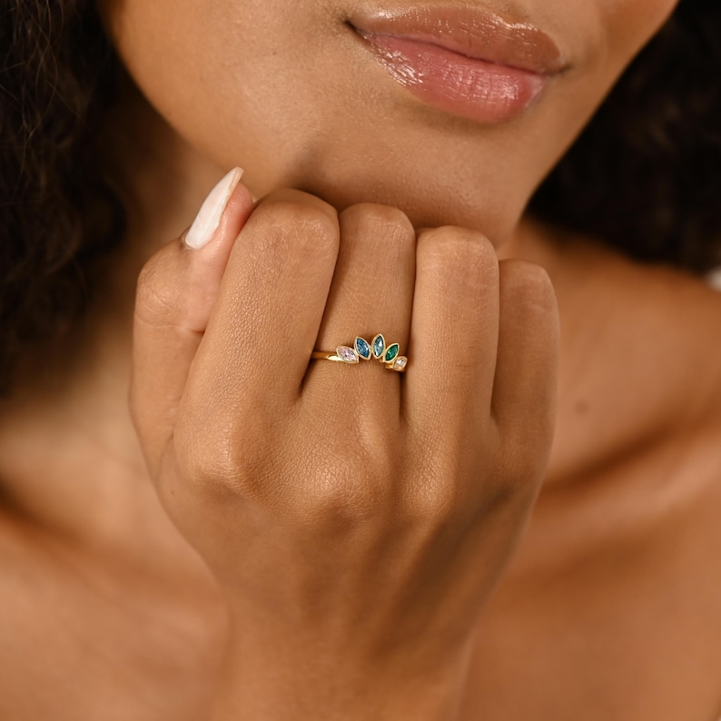 K&ouml;nnte beinhalten: Ein Goldring mit einer geschwungenen Anordnung von f&uuml;nf marquise-geschliffenen Edelsteinen in verschiedenen Blau-, Gr&uuml;n- und klaren Wei&szlig;t&ouml;nen. Die Steine sind f&auml;cherf&ouml;rmig auf der Oberseite des d&uuml;nnen, polierten Goldmetallrings angebracht.