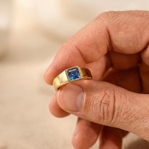 May include: A gold signet ring featuring a square-cut blue gemstone set in the centre. The band is smooth and polished, with a thick, solid construction that tapers slightly towards the back. The blue stone has a faceted surface that reflects light.
