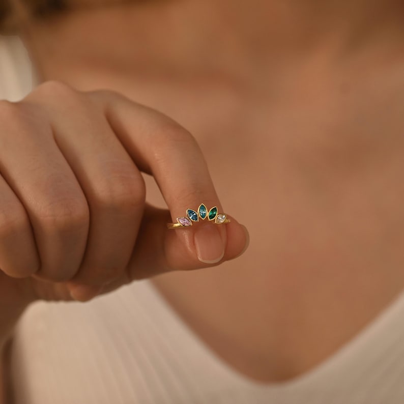 K&ouml;nnte beinhalten: Ein goldener Ring mit einer geschwungenen, kronenartigen Anordnung aus f&uuml;nf marquise-geschliffenen Edelsteinen in einem Farbverlauf aus Rosa, Hellblau, Dunkelblau, Gr&uuml;n und klarem Wei&szlig;, gefasst in einem zierlichen Metallrahmen.