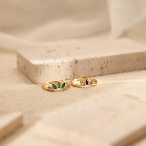 K&ouml;nnte beinhalten: Zwei goldene Ringe mit marquise-geschliffenen Steinen in F&auml;cherform. Der linke Ring hat gr&uuml;ne und helle Steine, w&auml;hrend der rechte Ring einen zentralen roten Stein, flankiert von hellen Steinen, auf einem d&uuml;nnen, polierten Goldband aufweist.