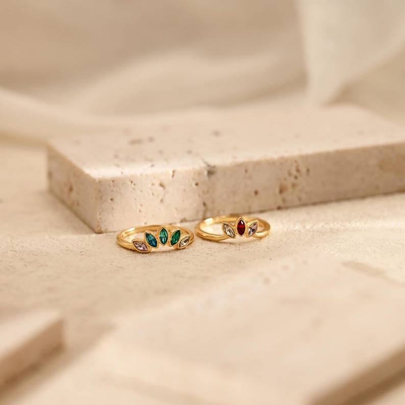 K&ouml;nnte beinhalten: Zwei goldene Ringe mit marquise-geschliffenen Steinen in F&auml;cherform. Der linke Ring hat gr&uuml;ne und helle Steine, w&auml;hrend der rechte Ring einen zentralen roten Stein, flankiert von hellen Steinen, auf einem d&uuml;nnen, polierten Goldband aufweist.