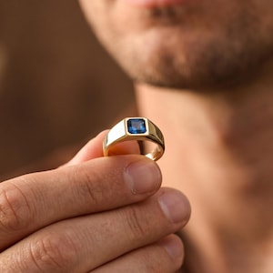 May include: A gold signet ring featuring a square-cut blue gemstone set in the centre. The band is polished and smooth, with a flat top surface that holds the vibrant blue stone securely in place, creating a classic and refined jewellery piece.