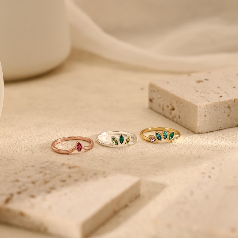 K&ouml;nnte beinhalten: Drei Ringe in einer Reihe angeordnet. Der erste ist ein Ros&eacute;goldring mit einem einzelnen rosafarbenen Stein im Marquise-Schliff. Der zweite ist ein Silberring mit drei gr&uuml;nen Steinen im Marquise-Schliff. Der dritte ist ein Goldring mit f&uuml;nf bunten Steinen im Marquise-Schliff.