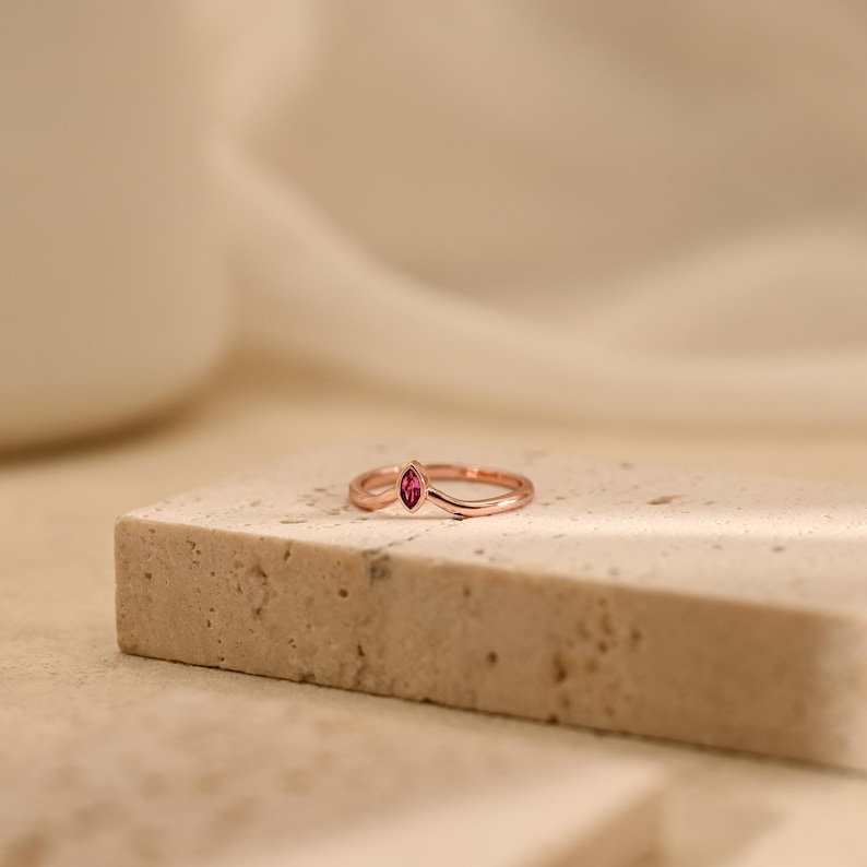 K&ouml;nnte beinhalten: Ein Ring aus Ros&eacute;gold mit einem V-f&ouml;rmigen Band und einem einzelnen, im Marquise-Schliff geschliffenen rosa Edelstein in der Mitte. Das Metallband hat eine glatte, polierte Oberfl&auml;che und der Edelstein sitzt an der Spitze des Chevron-Designs.