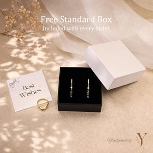 K&ouml;nnte beinhalten: Ein Goldring mit einem einzelnen Stein, ein Paar Ohrh&auml;nger mit kleinen blauen und klaren Steinen sowie eine wei&szlig;e Geschenkbox. Eine Karte mit dem Text Best Wishes liegt bei. Der Text Free Standard Box Included with every order ist zu sehen.
