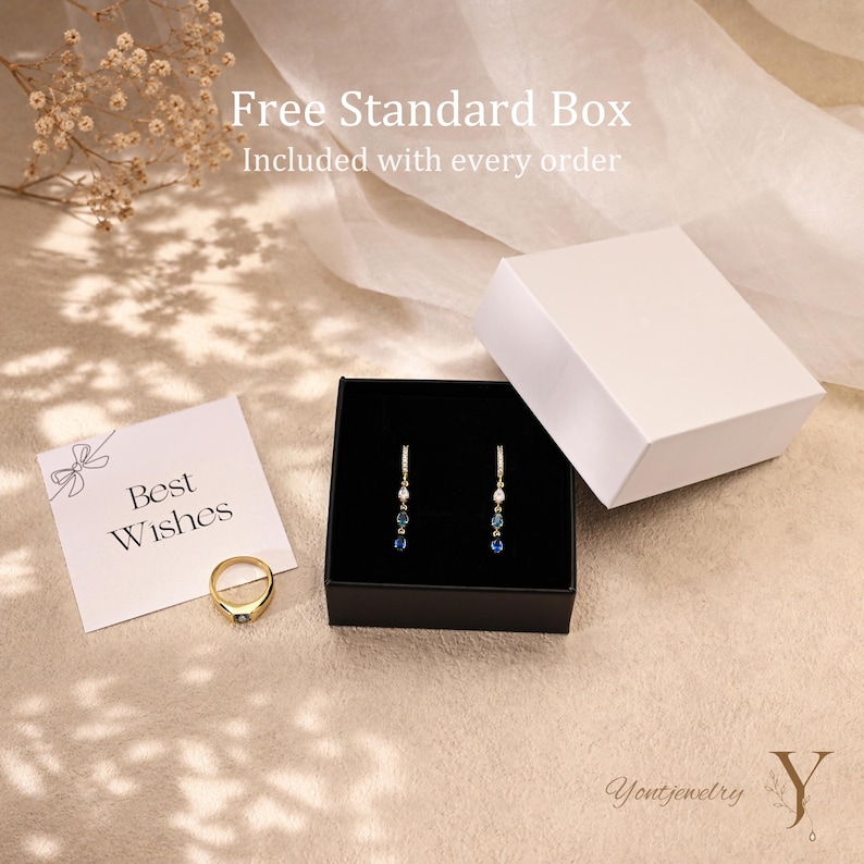 K&ouml;nnte beinhalten: Ein Goldring mit einem einzelnen Stein, ein Paar Ohrh&auml;nger mit kleinen blauen und klaren Steinen sowie eine wei&szlig;e Geschenkbox. Eine Karte mit dem Text Best Wishes liegt bei. Der Text Free Standard Box Included with every order ist zu sehen.