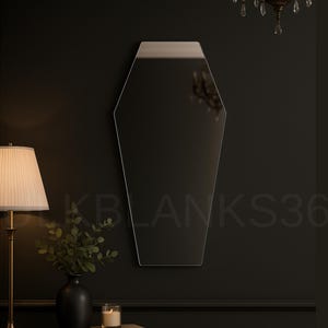 May include: A coffin-shaped mirror with a silver top edge hangs on a dark wall. The mirror reflects the room's details. A lamp with a beige lampshade and a black vase with greenery sit on a dark table. A candle and a book are also on the table.