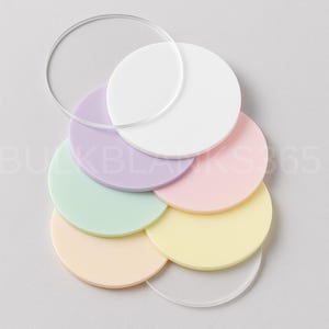 May include: A collection of colorful, round acrylic blanks in pastel shades of white, lavender, pink, yellow, mint green, and peach. Two clear acrylic circles are also included. The image is taken from an overhead perspective.