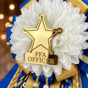 May include: A gold star-shaped FFA Officer badge with the text "FFA OFFICER" on a white flower, accented with blue and gold ribbons. The badge is a decorative item, likely for a school or organization.