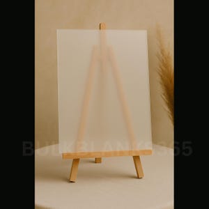 May include: A small wooden easel with a frosted acrylic panel. The easel is made of light-colored wood and has a simple A-frame design. The acrylic panel is rectangular and appears translucent. The text "BULKBLANKS365" is printed at the bottom.