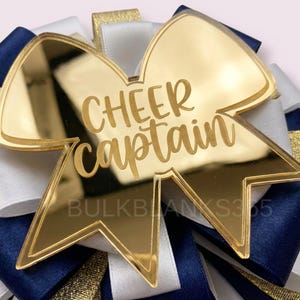 May include: A gold-colored acrylic bow with the words "CHEER captain" in a script font. The bow is layered over navy blue, white, and gold ribbons. The text "BULKBLANKS365" is at the bottom.