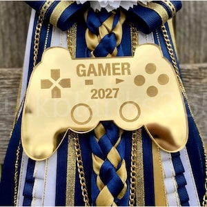 May include: A decorative homecoming mum featuring a gold-toned acrylic video game controller cutout engraved with the text GAMER 2027. The accessory is adorned with layered navy blue, white, and gold ribbons, braided fabric accents, and metallic gold chains.