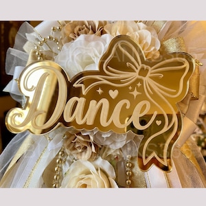 May include: A gold mirror acrylic cutout featuring the word Dance in cursive script, accented with an engraved bow, hearts, and stars. The cutout is layered over a decorative arrangement of white and beige fabric flowers, tulle, and gold ribbon.