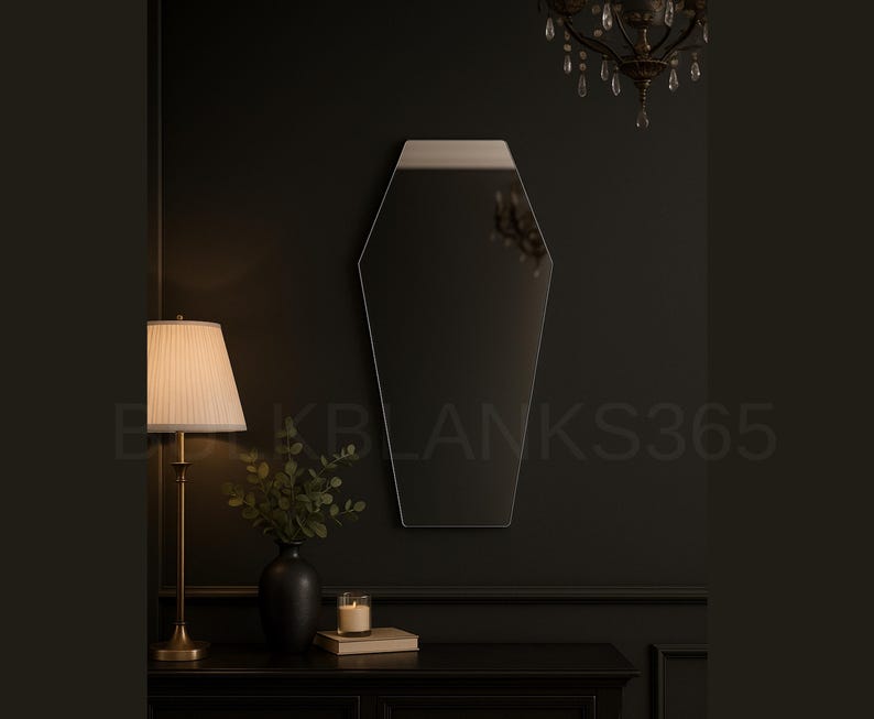 K&ouml;nnte beinhalten: Ein Sarg-f&ouml;rmiger Spiegel mit silberner Oberkante h&auml;ngt an einer dunklen Wand. Der Spiegel reflektiert die Details des Raumes. Eine Lampe mit beigem Lampenschirm und eine schwarze Vase mit Gr&uuml;n stehen auf einem dunklen Tisch.