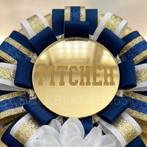 May include: A gold-colored rosette with navy blue, white, and gold ribbons. The center features the word "PITCHER" in a bold font. The rosette is a decorative item, possibly for sports or celebrations.