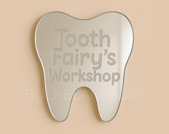 May include: A silver-colored tooth-shaped sign with the words "Tooth Fairy's Workshop" engraved on it. The sign is set against a light beige background. The sign is a decorative item.