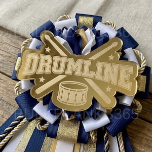 May include: A decorative rosette badge featuring navy blue and white ribbon loops with gold trim. A central gold acrylic plaque displays the text DRUMLINE, a graphic of two crossed drumsticks, four small stars, and a snare drum illustration.