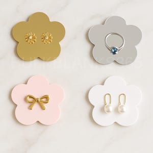 May include: Four flower-shaped jewelry holders in gold, silver, pink, and white. Each holds different jewelry: daisy earrings, a ring with a blue stone, a gold bow, and pearl earrings. The holders are on a marble surface.