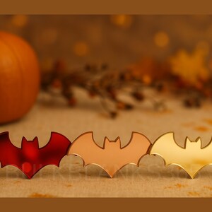 May include: Three bat-shaped decorations in red, rose gold, and gold. The bats are arranged in a row, with a pumpkin and fall foliage in the background. The bats are likely for Halloween or fall decor.
