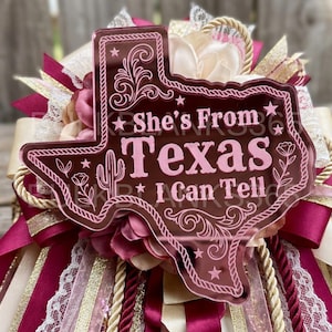 May include: A Texas-shaped maroon acrylic plaque featuring the text She's From Texas I Can Tell, accented with stars, floral motifs, and a cactus. The plaque is layered over a decorative arrangement of maroon and cream ribbons, lace, and rope.
