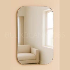 May include: A rectangular mirror with rounded corners hangs on a beige wall. The mirror's reflection shows a room with a window, a beige armchair, and a curtain. The mirror has a thin, dark frame. The text "BULKBLANK365" is visible in the image.