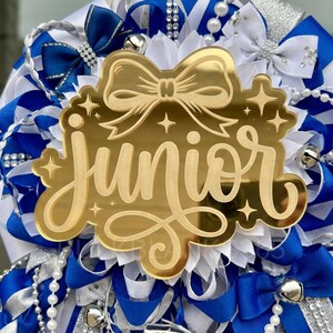 May include: A gold-colored acrylic cutout with the word "Junior" in elegant script, surrounded by star accents and a bow design. The cutout is set against a backdrop of blue, white, and silver ribbons, bows, and decorative elements, including small bells and beaded strands.