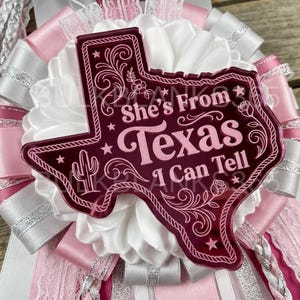 May include: A decorative rosette badge featuring a dark pink acrylic Texas-shaped plaque with the text She's From Texas I Can Tell, a cactus, and floral engravings. The plaque is layered over a ruffled white satin ribbon base with pink and silver accents.