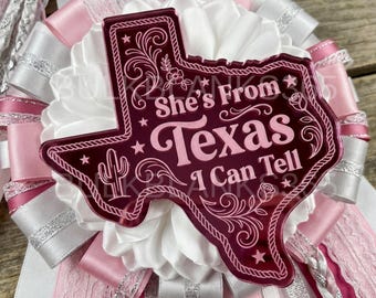 May include: A decorative rosette badge featuring a dark pink acrylic Texas-shaped plaque with the text She's From Texas I Can Tell, a cactus, and floral engravings. The plaque is layered over a ruffled white satin ribbon base with pink and silver accents.