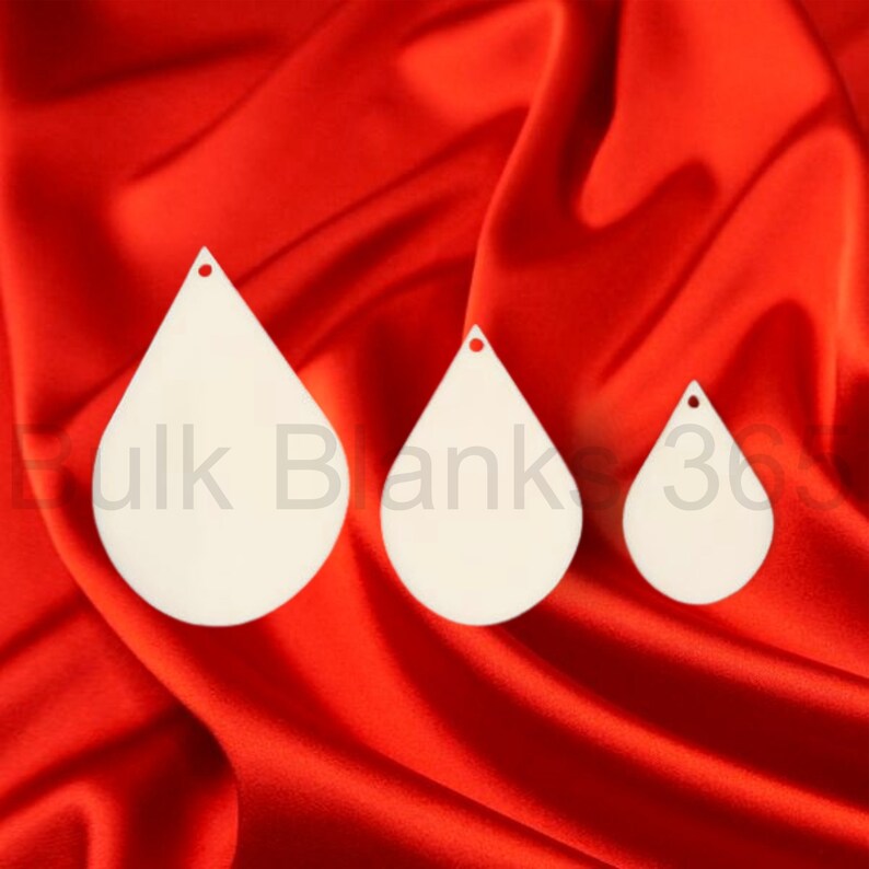 May include: Three white teardrop-shaped wood blanks with a hole at the top. The blanks are arranged in descending size order from left to right. The blanks are on a red fabric background.