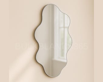 May include: An irregularly shaped wall mirror with a silver frame. The mirror has a wavy, organic design, and is mounted on a light beige wall. The reflection shows a window and natural light.