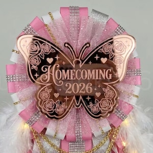 May include: A homecoming mum badge featuring a rose gold acrylic butterfly centerpiece engraved with the text HOMECOMING 2026 and rose floral patterns. The badge is surrounded by layers of pink and white ribbon, silver glitter accents, rhinestones, gold chains, and white feathers.
