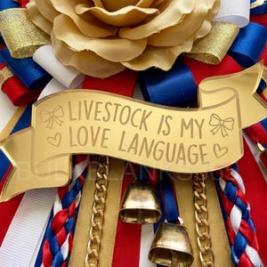 May include: A gold-colored banner with the text "Livestock is my love language" is the focal point of this image. The banner is surrounded by red, white, and blue ribbons, gold chains, and small gold bells. A large, tan flower is at the top.