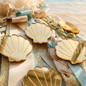 May include: Close-up of several gold-colored seashell-shaped decorations. The shells are arranged on a bed of ribbons in shades of blue, gold, and cream. The image has a soft, beachy aesthetic.