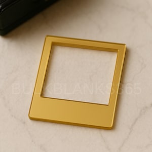 May include: A gold-colored acrylic blank in the shape of a vintage instant photo frame. The frame has a rectangular shape with a central opening. The surface is smooth and reflective. The item is on a light-colored surface.