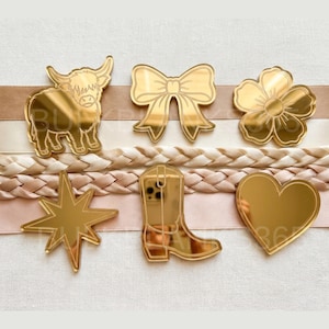 May include: A collection of gold-colored acrylic charms in various shapes, including a Highland cow, a bow, a flower, a star, a cowboy boot, and a heart. The charms are arranged on a background of ribbons and braided trim.