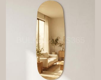 May include: An oval-shaped, frameless mirror with a warm, golden hue hangs on a white wall. The mirror reflects a living room scene with a white sofa, wooden furniture, and natural light. The mirror's elongated shape adds a modern touch to the space.