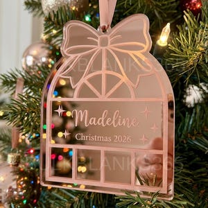 May include: A rose gold mirror acrylic Christmas ornament shaped like a window with a bow at the top. The ornament is engraved with the name Madeline and the text Christmas 2026, featuring four rectangular panels at the bottom and a ribbon hanger.