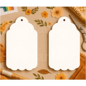 May include: Two blank, off-white gift tags with a scalloped bottom edge and a hole at the top. The tags are on a tan surface, surrounded by floral and craft supplies, including scissors and a marker.