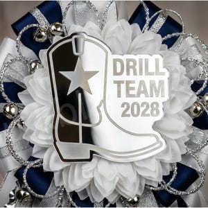 May include: A decorative item featuring a silver cowboy boot with a star, the words "DRILL TEAM 2028", white fabric, navy blue and silver ribbons, and silver bells. The item has a layered design with a mix of textures and colors.