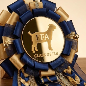May include: A decorative FFA ribbon in gold, navy blue, and gold. The center features a gold disc with a goat silhouette and the letters "FFA" and the text "CLASS OF '28". The ribbon is made of layered satin and sheer ribbons.
